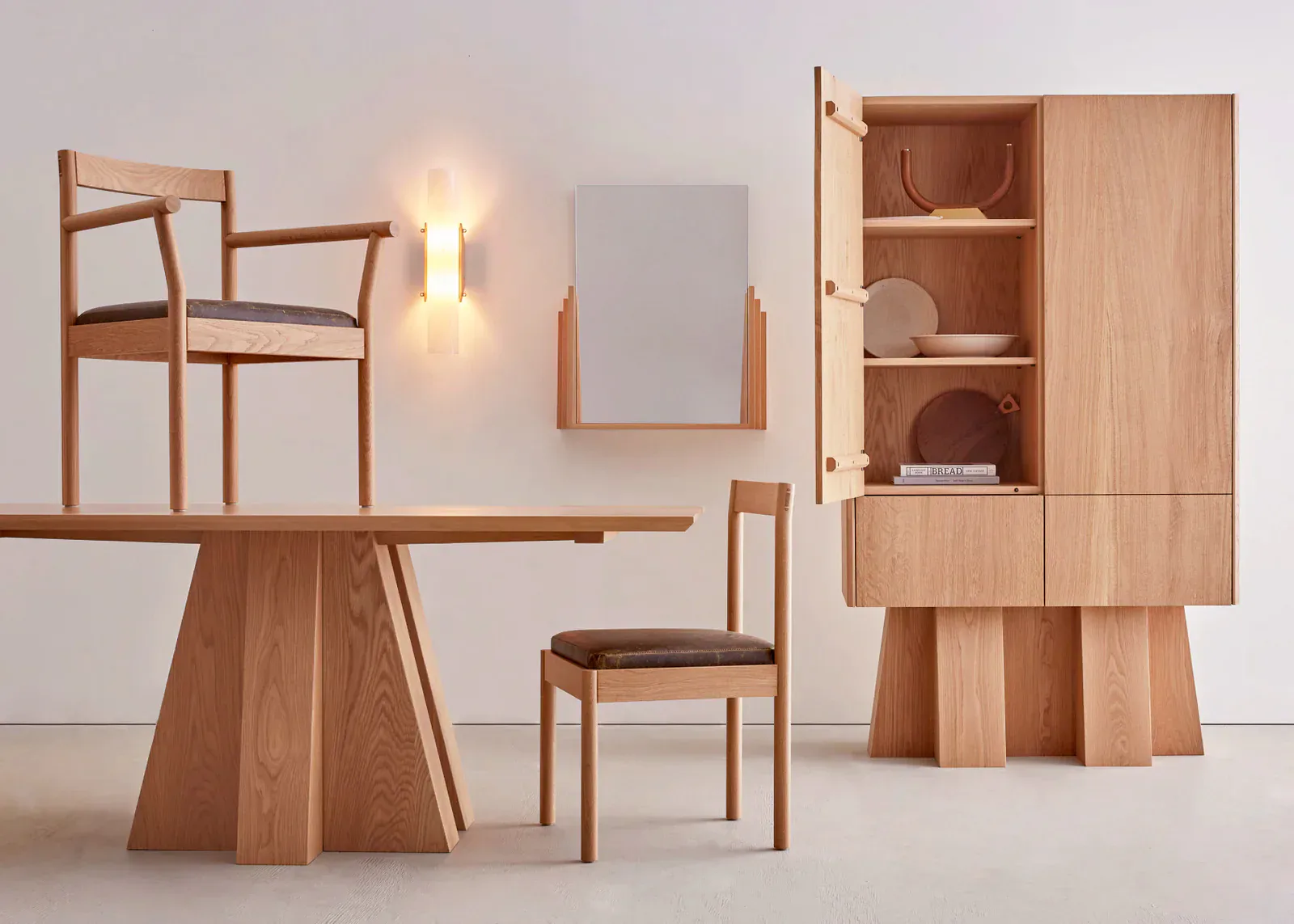 Wooden furniture collection featuring a dining table with geometric base, chairs, wall-mounted shelving unit, and accent lighting in natural wood tones by Bowen Liu Studio.