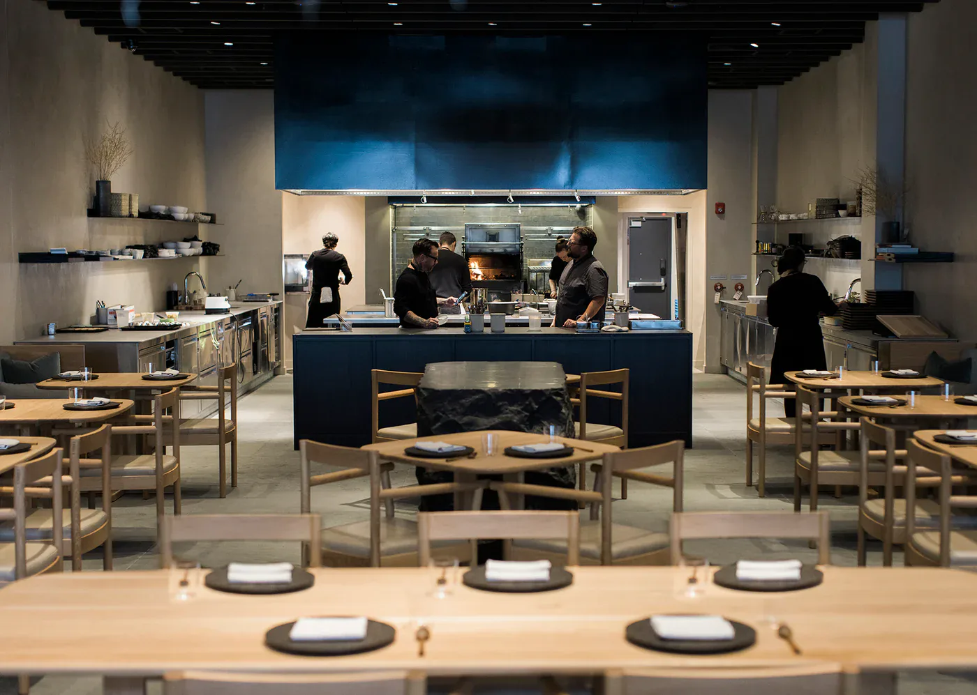 Modern restaurant interior with open kitchen, blue accent wall, wooden dining chairs, and counter seating by Bowen Liu Studio.