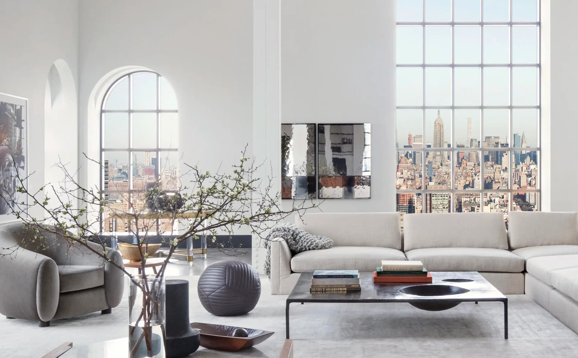 Modern living room with floor-to-ceiling windows overlooking a city skyline, featuring a light gray sectional sofa, minimalist coffee table, and sculptural decor by Brad Ford ID.