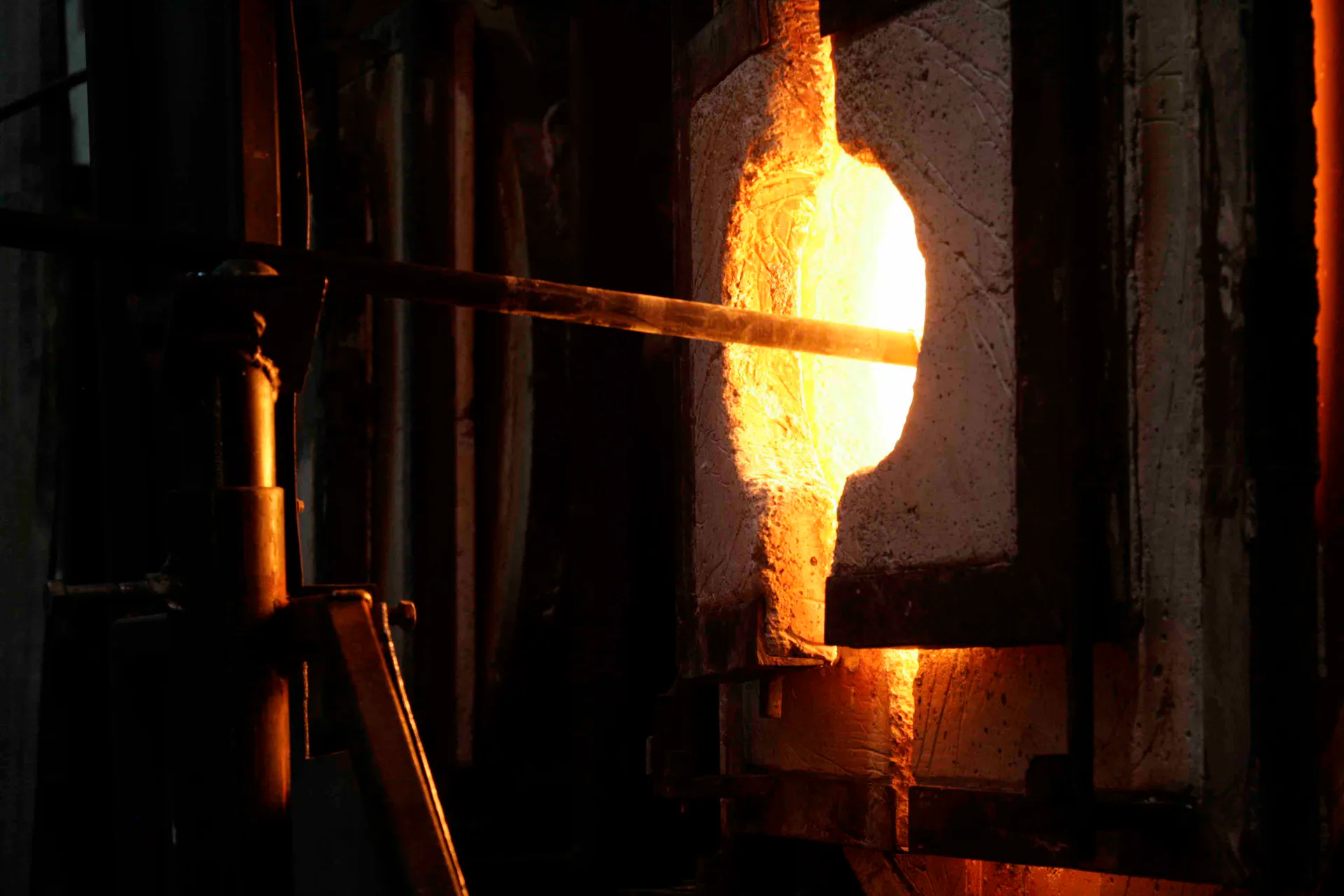 Glowing molten glass sphere at furnace opening with metal rod, showcasing the glassblowing process and heat of the kiln by Brooklyn Glass.