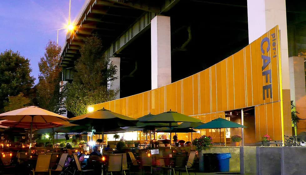 Evening view of a modern outdoor pavilion with golden corrugated metal cladding and integrated lighting beneath a concrete overpass, featuring dining areas and landscaping by Buck Moorhead Architect.