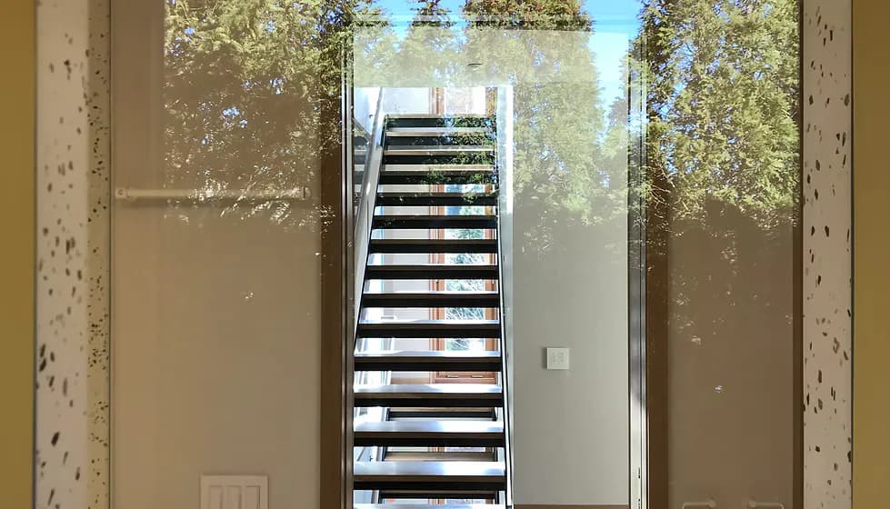 Modern staircase with black and white striped steps viewed through glass doors, flanked by concrete walls and framing views of trees, by Buck Moorhead Architect.