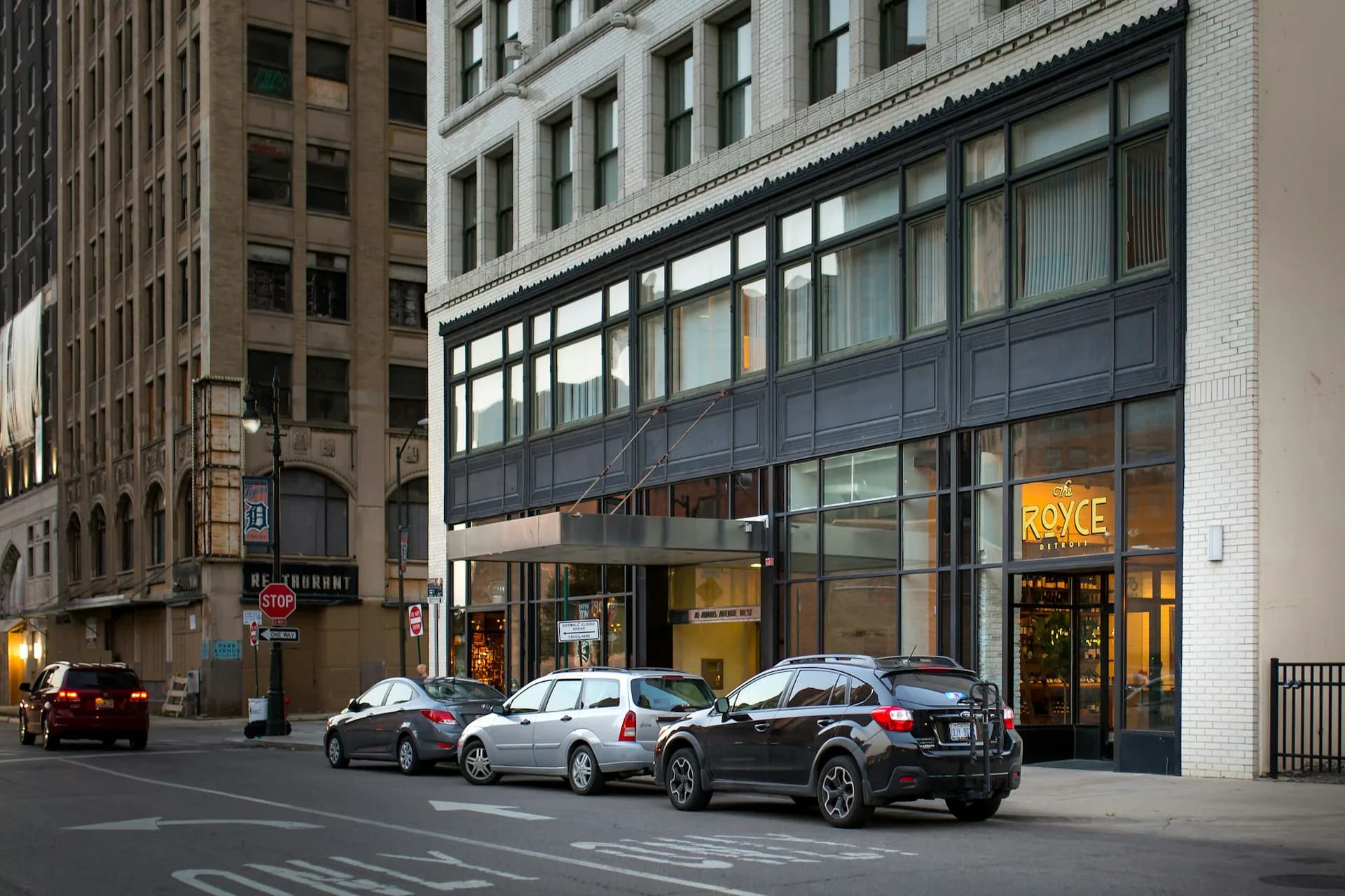 Modern street-level storefront with dark metal and glass facade beneath historic brick office building, featuring lit interior retail space and parked vehicles, designed by BWArchitects.