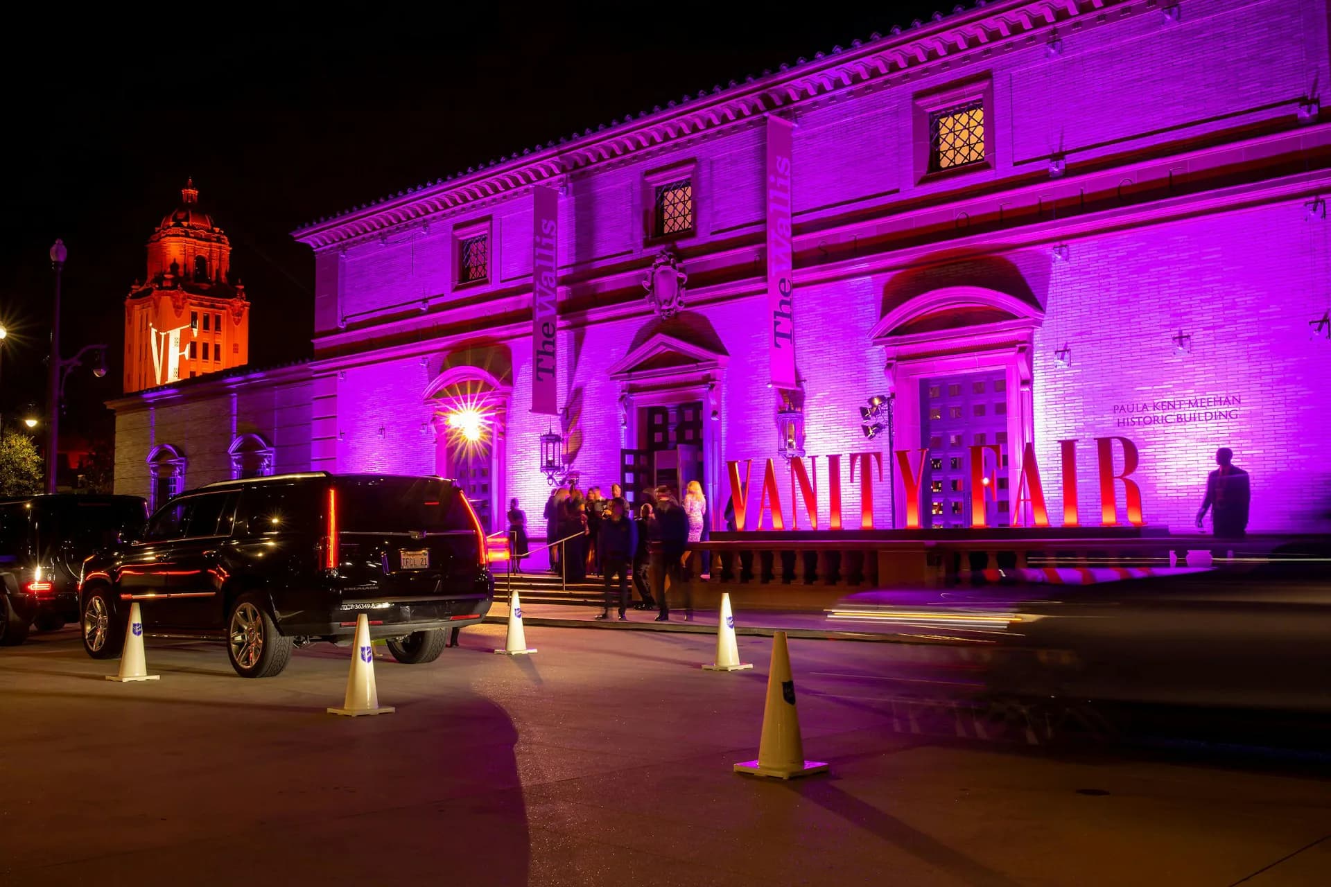 Exterior nighttime photograph of a neoclassical building facade illuminated in vibrant purple and magenta lighting with signage, featuring columned entrance and refined architectural details by BWArchitects.
