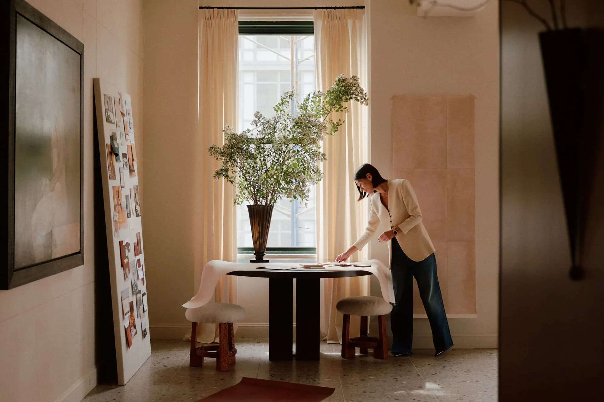 A warm, minimalist interior with peachy-toned walls features a black round table with potted greenery by a tall window with sheer curtains, styled by Calico Wallpaper.