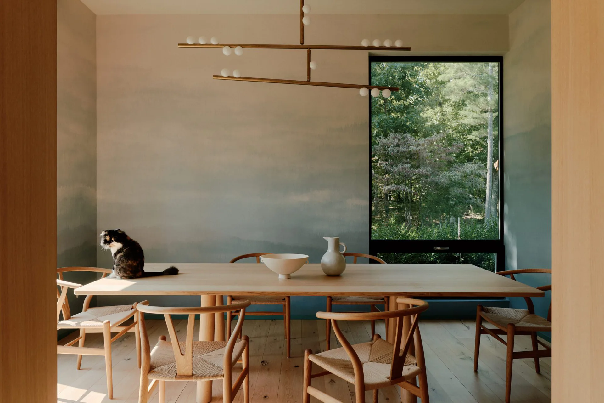 Minimalist dining room with natural wood chairs and table, warm plaster walls, pendant lighting, and large window overlooking greenery by Calico Wallpaper.