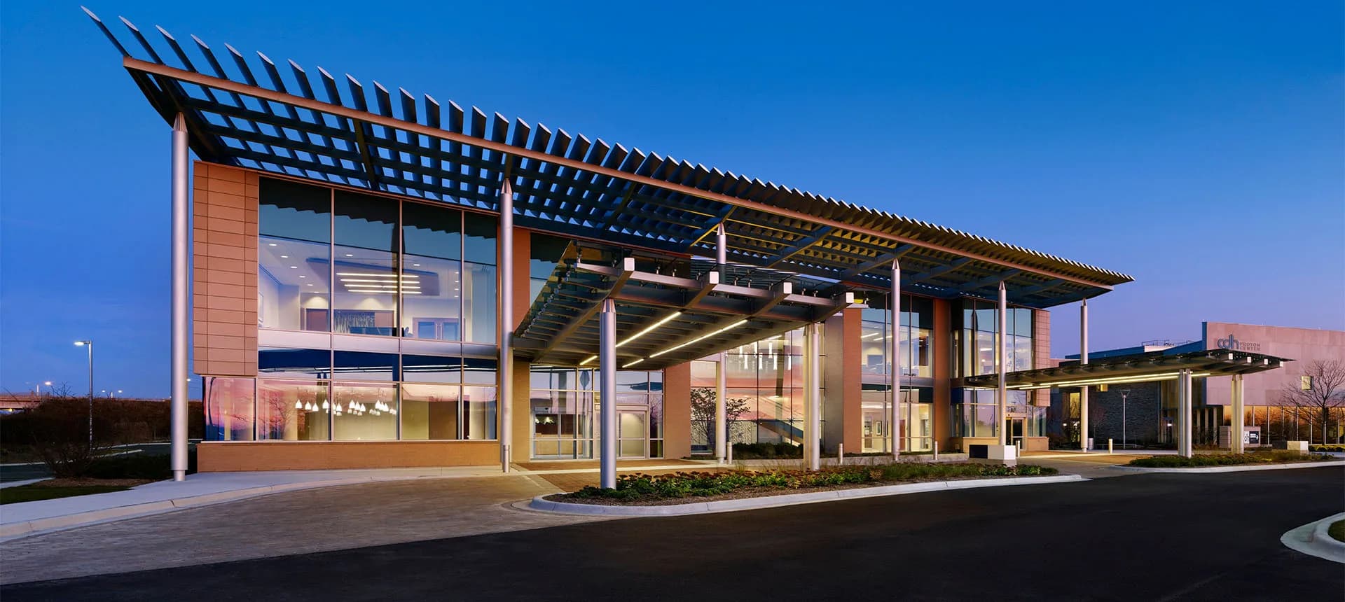 Modern institutional building with warm brick facade, glass curtain wall, and angular pergola structure illuminated at dusk by CallisonRTKL.