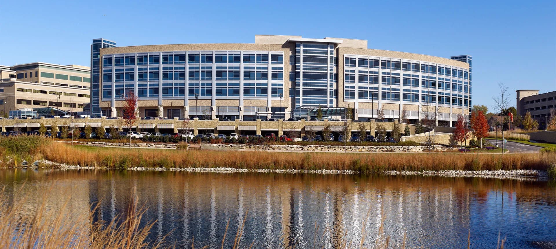 Modern multi-story office building with glass and stone façade reflected in waterfront pond, surrounded by landscaping, by CallisonRTKL.