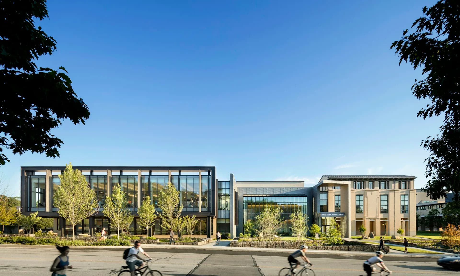 Modern institutional building with glass and brick facades, landscaped plaza with trees and cyclists, clear sky, by CannonDesign.
