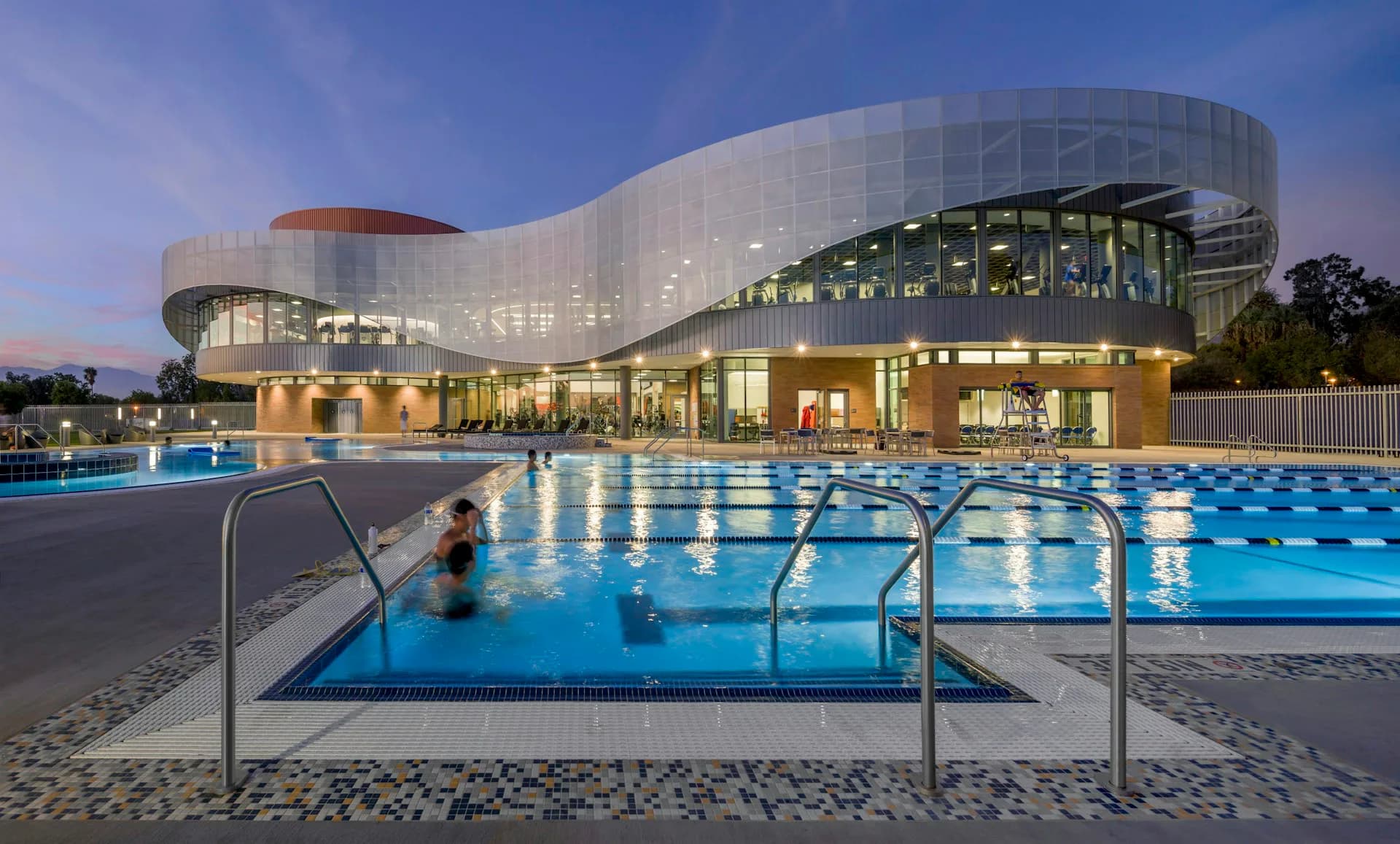 Modern aquatic center with curved white facade, illuminated at dusk, featuring Olympic-length pool with swimmers and diving boards by CannonDesign.