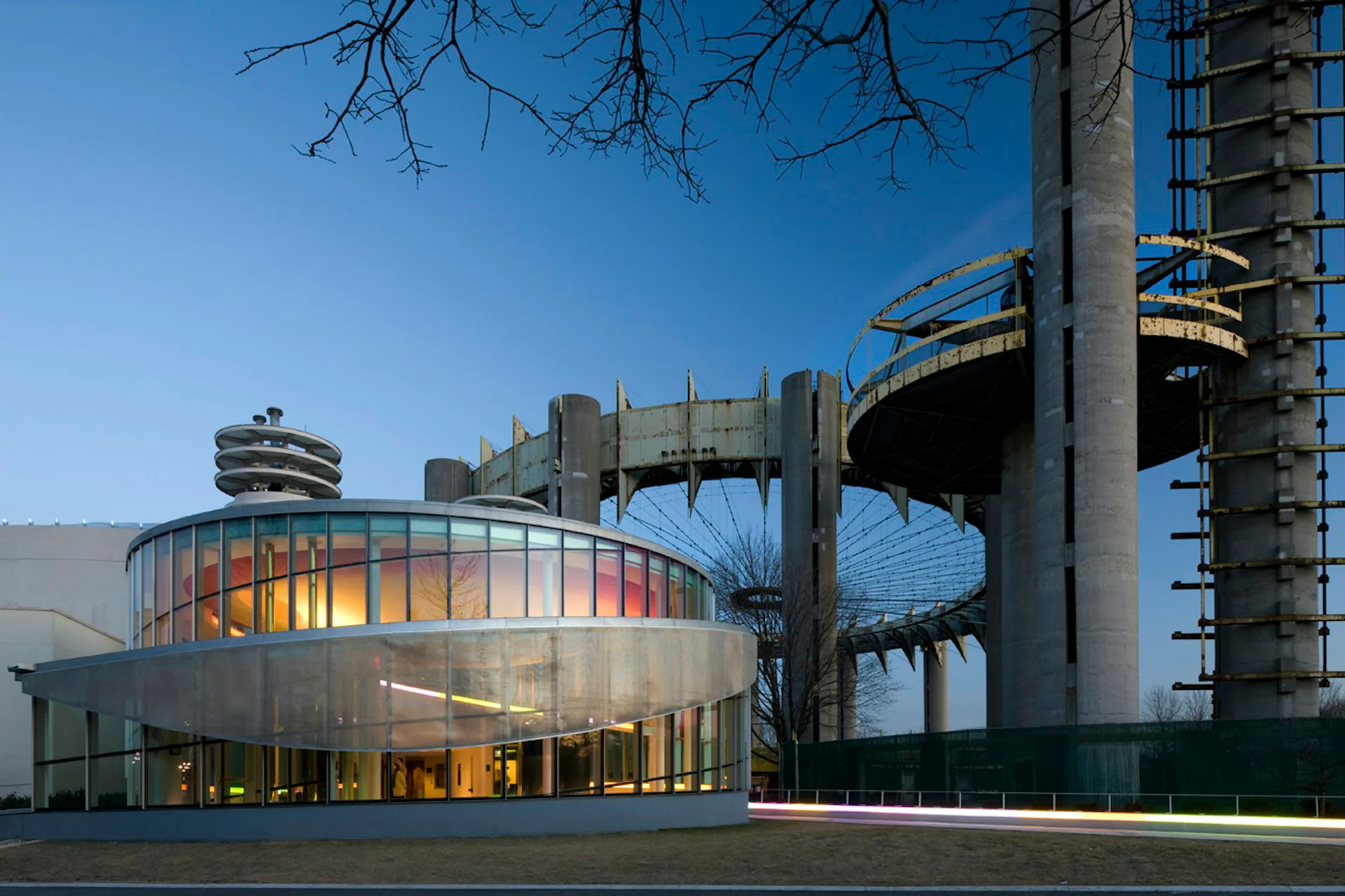 Curved modernist building with illuminated glass facade and cylindrical tower element set against brutalist concrete structure by Caples Jefferson Architects.