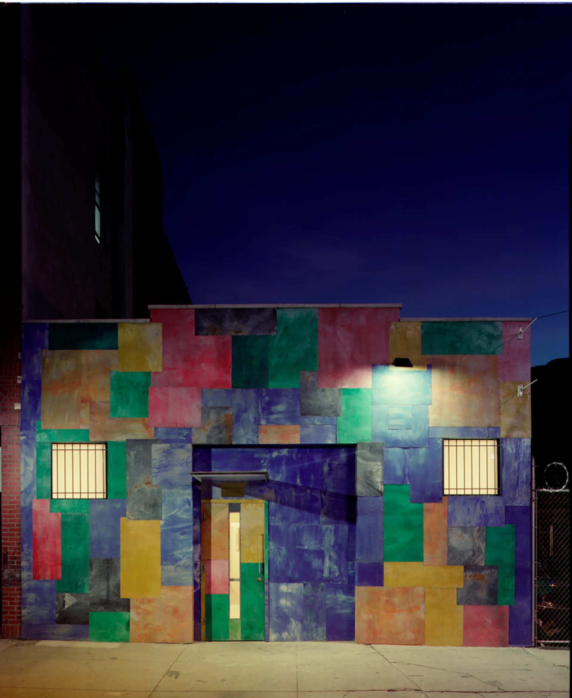 Nighttime photograph of a colorful facade with irregularly arranged colored panels in red, green, blue, yellow, and purple, featuring two illuminated window grilles, designed by Caples Jefferson Architects.