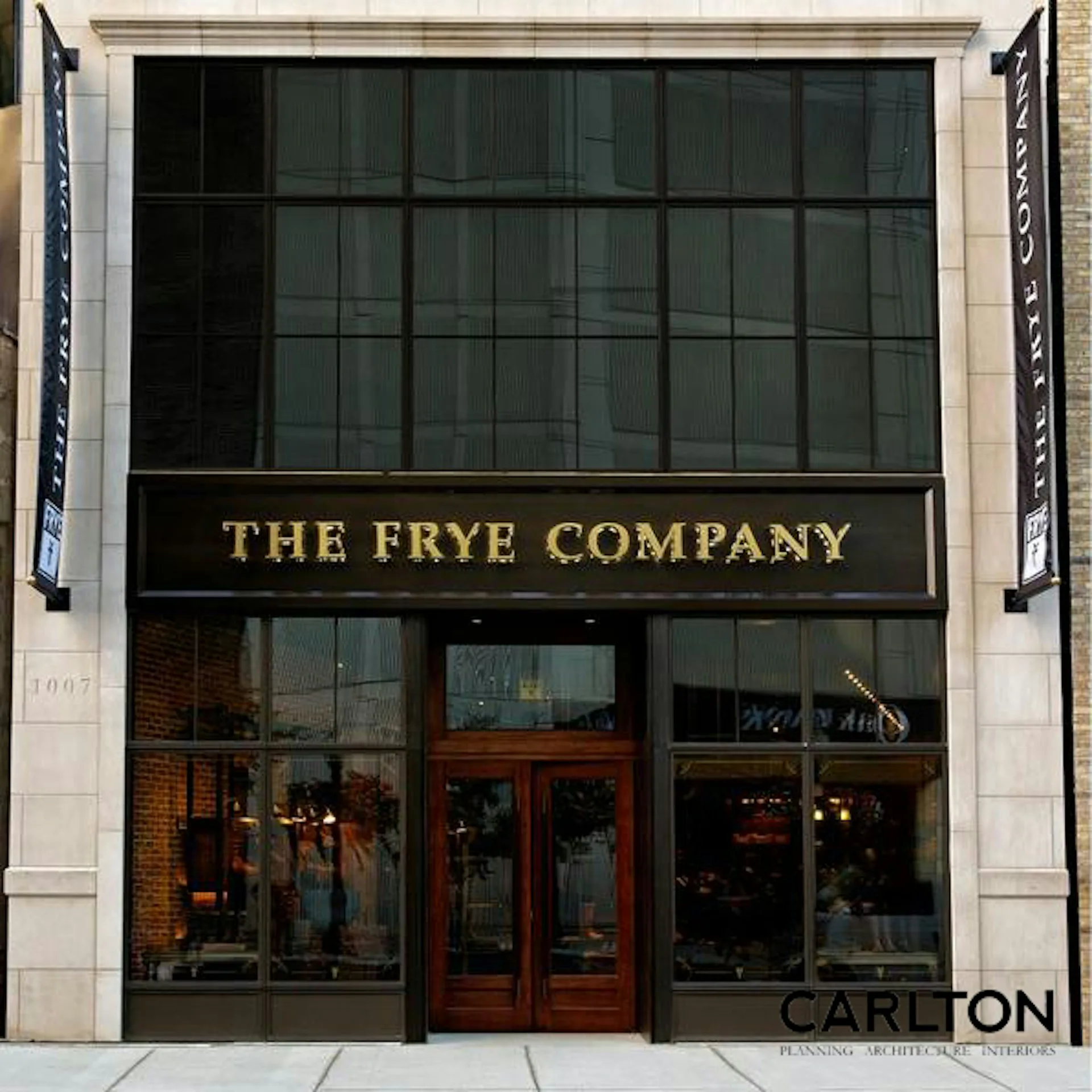 The Frye Company storefront featuring large gridded windows, red entry doors, and gold lettering on a dark band, designed by Carlton Architecture PC.