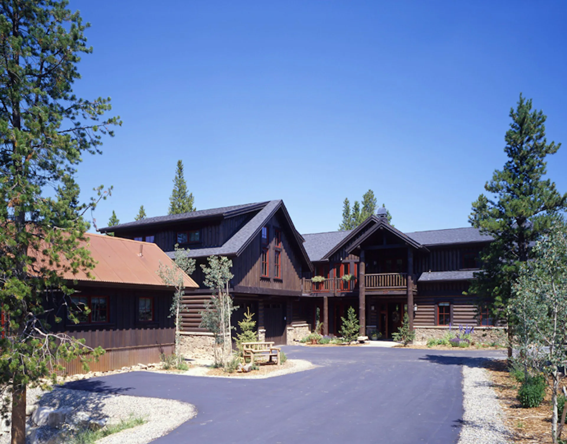 Large residential lodge with dark wood siding and pitched rooflines nestled among evergreen trees, featuring a curved driveway and natural stone landscaping by Carol Maryan Architects, PC.