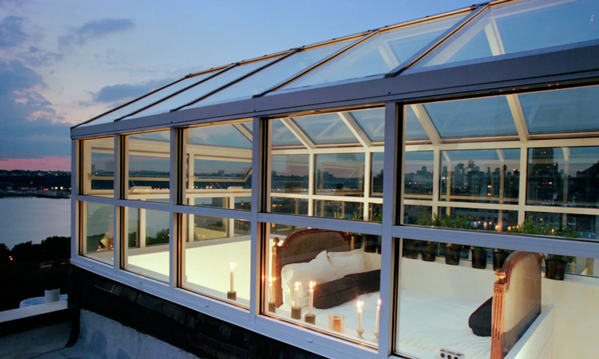 Modern glass-enclosed rooftop room with angular skylight framing views of waterfront and distant shoreline at dusk, featuring warm interior lighting and minimalist furnishings by Carol Maryan Architects, PC.