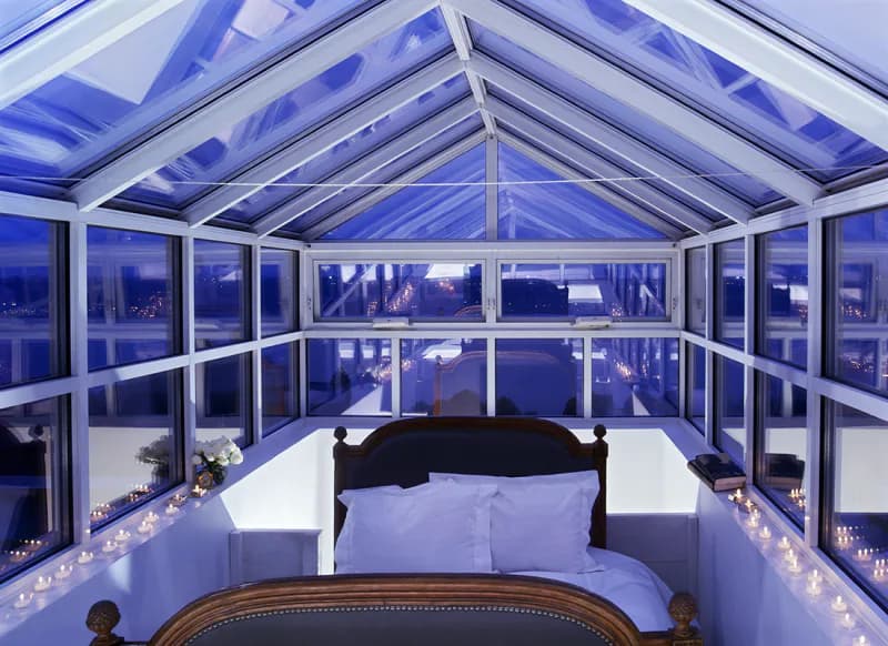 Modern glass conservatory with blue-tinted ceiling panels and white framing, featuring panoramic windows overlooking a nighttime cityscape, designed by Carol Maryan Architects, PC.
