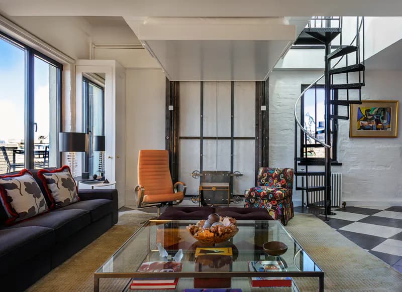 Contemporary loft living room featuring a dark sofa, orange accent chair, glass coffee table, and industrial spiral staircase, with large windows and minimalist design by Carol Maryan Architects, PC.