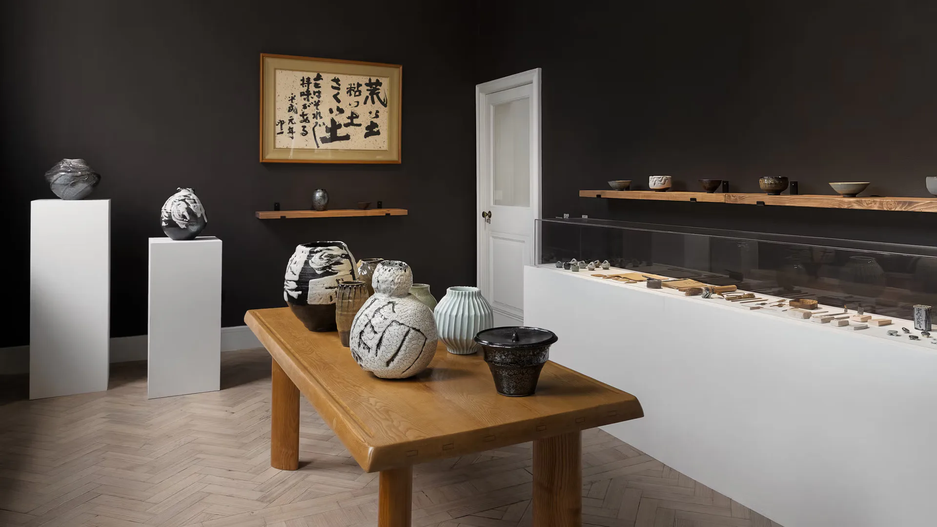 Dark gallery interior with wooden display table, white pedestals, ceramic vessels, and Japanese calligraphy artwork by Carpenters Workshop Gallery.