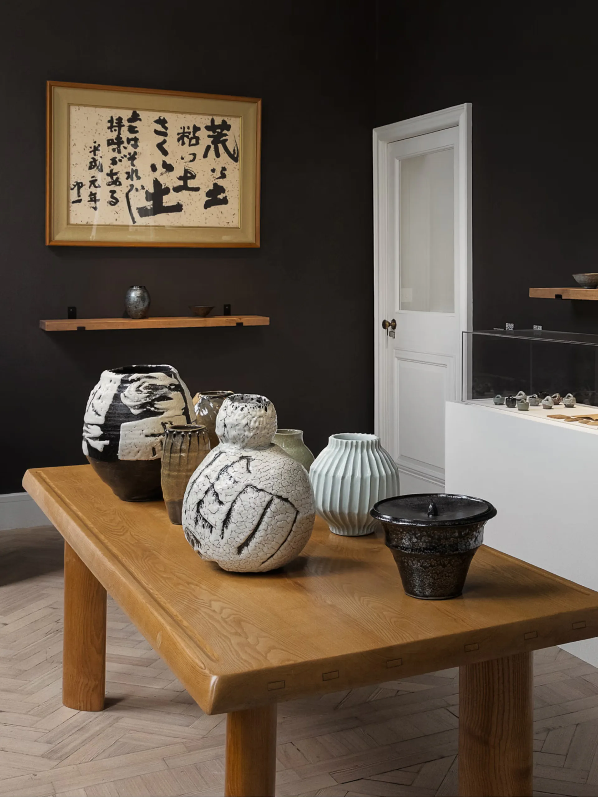 Dark gallery interior with wooden table displaying ceramic vessels and Asian art, white door visible, framed calligraphy on wall by Carpenters Workshop Gallery.