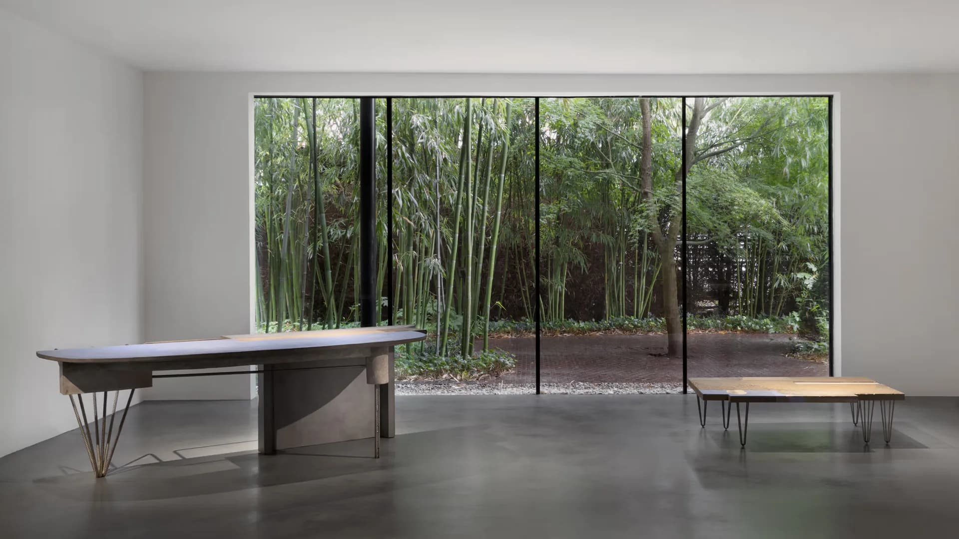 Minimalist interior with dark sculptural furniture and polished concrete floor overlooking a bamboo forest through floor-to-ceiling glass by Carpenters Workshop Gallery.
