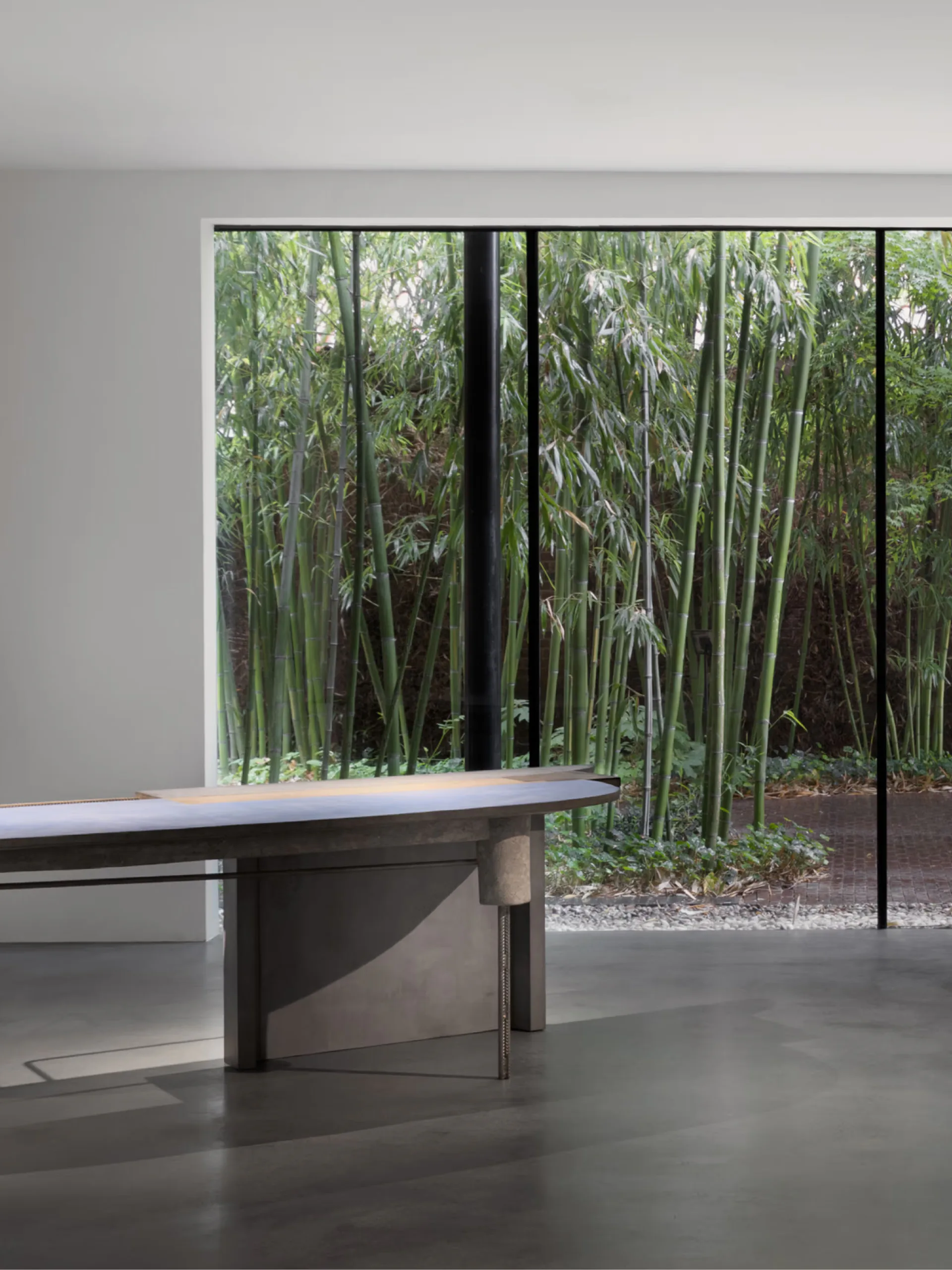 Minimalist interior with a geometric dining table overlooking a dense bamboo grove through floor-to-ceiling windows by Carpenters Workshop Gallery.