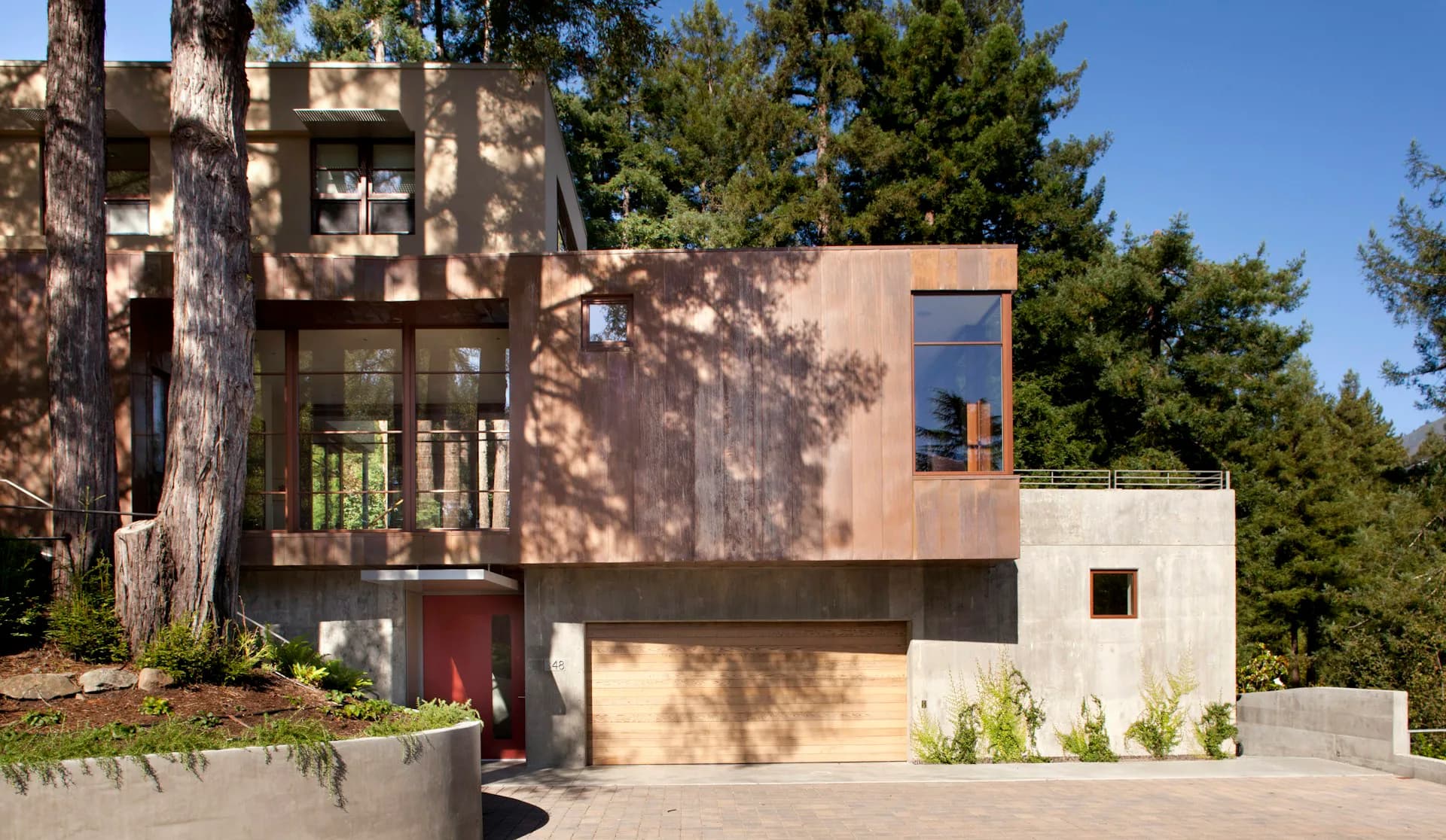 Modern residential exterior featuring mixed materials including copper cladding, concrete, and natural wood siding nestled among tall evergreen trees by Cass Calder Smith Architecture + Interiors.