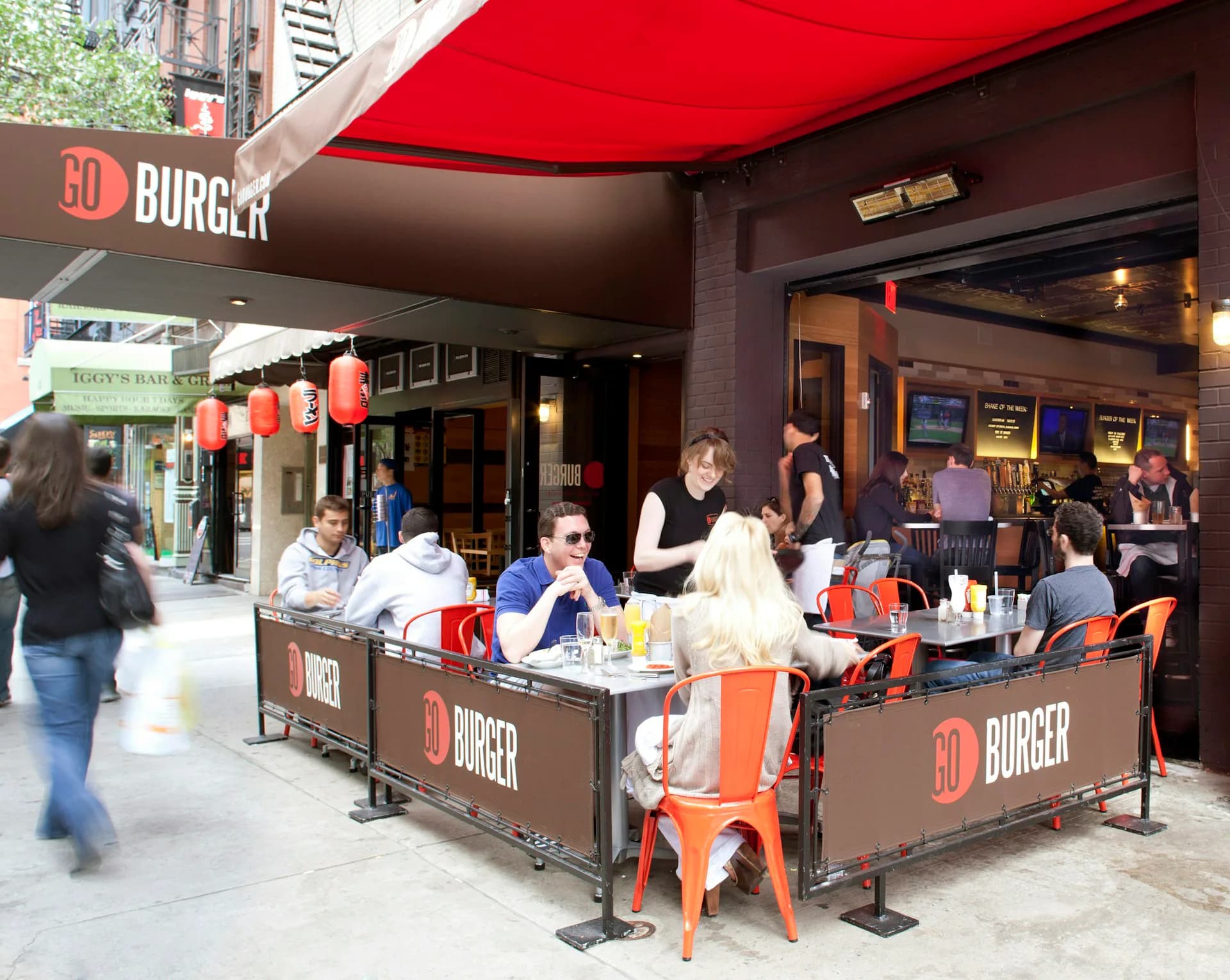 Outdoor burger restaurant with red awning, orange metal chairs, and branded signage featuring a lively streetside seating area by Cass Calder Smith Architecture + Interiors.