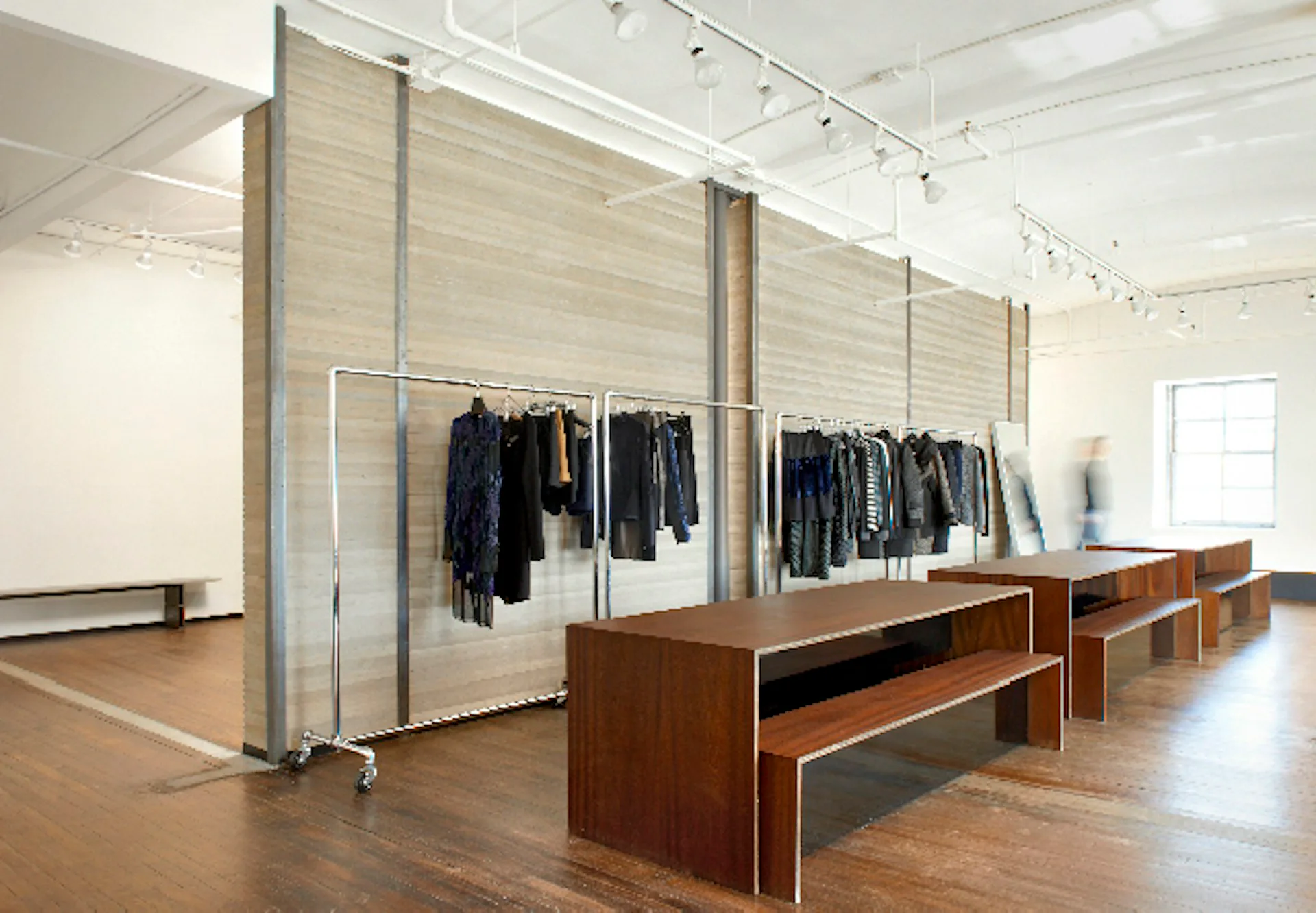 Modern retail showroom with wood-paneled partitions, clothing racks displaying dark garments, and wood benches, featuring polished floors and track lighting by CDR Studio Architects, PC.