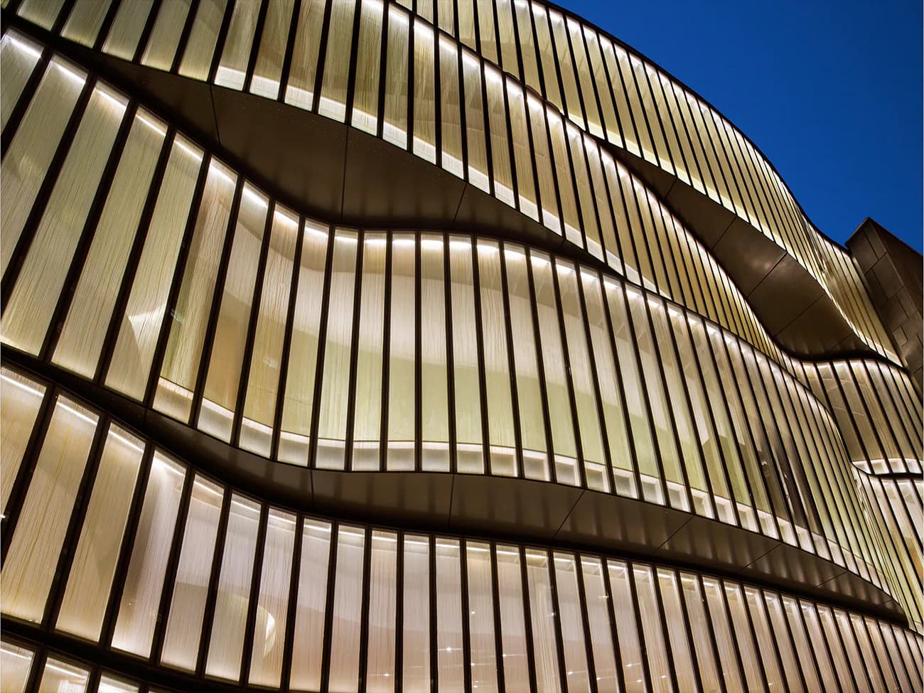 Illuminated facade with horizontal corrugated metal panels and curved architectural elements against deep blue sky by CetraRuddy.