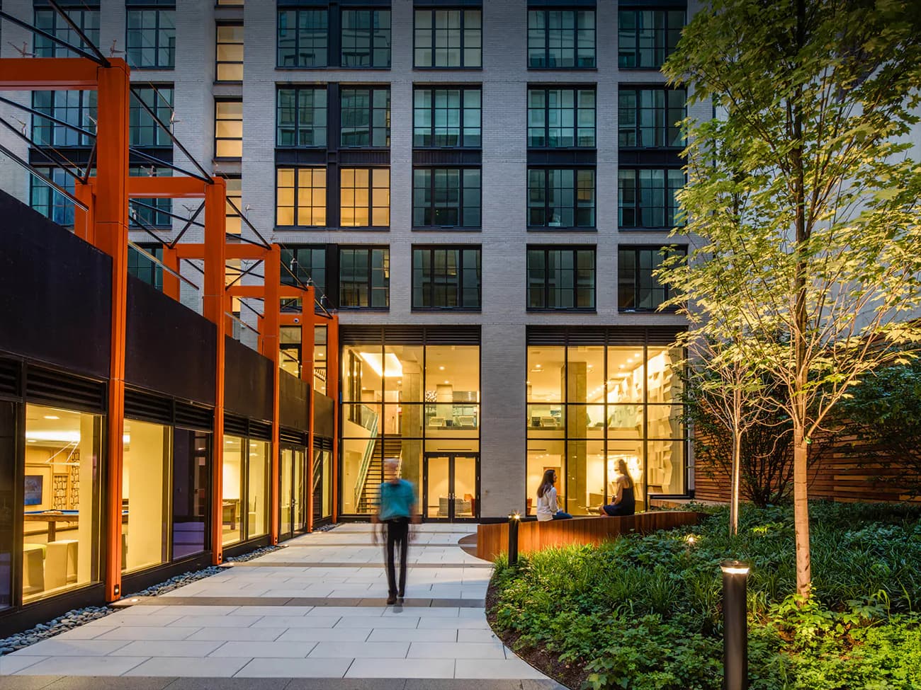 Evening courtyard with illuminated storefronts, orange steel pergola, planted trees, and modern residential tower above by CetraRuddy.