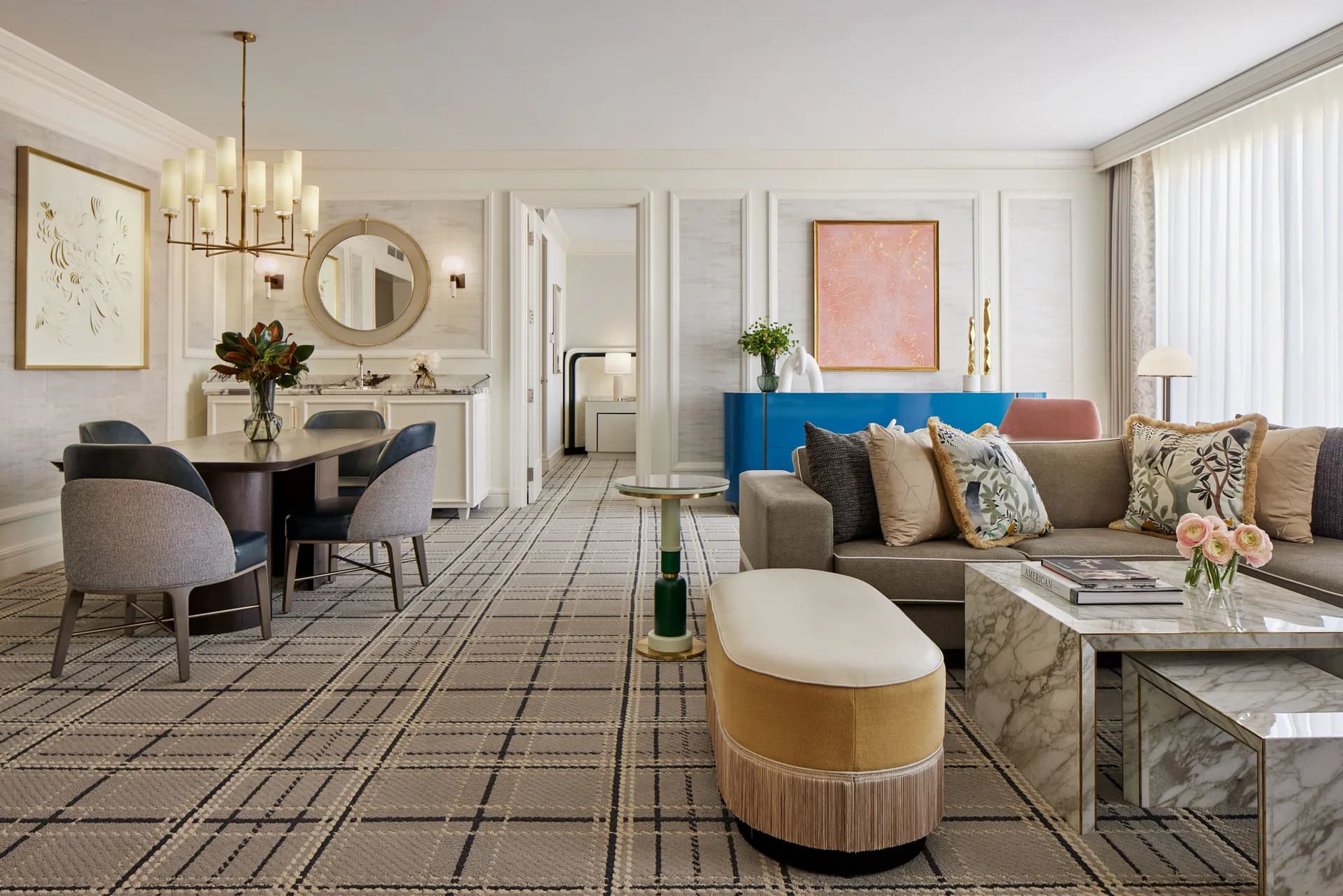 Luxurious hotel suite with gray upholstered sofa, gold-striped ottoman, marble side table, and brass accents, featuring soft neutral palette and geometric patterned flooring by Champalimaud Design.
