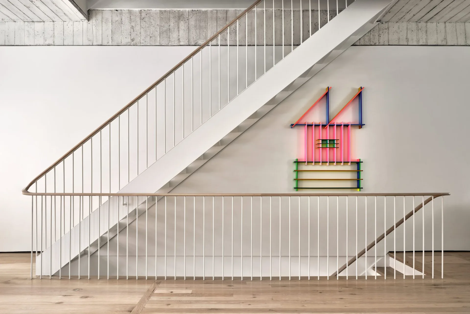 Minimalist interior staircase with metal railings and a colorful geometric house sculpture installation on white wall by Chango & Co.