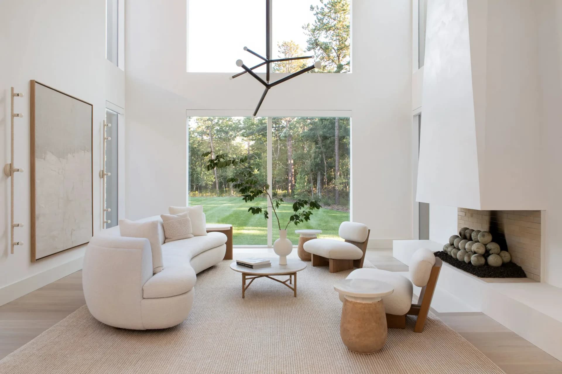 Bright modern living room with curved cream furniture, wood accents, large windows overlooking landscape, fireplace, and sculptural ceiling fixture by Chango & Co.