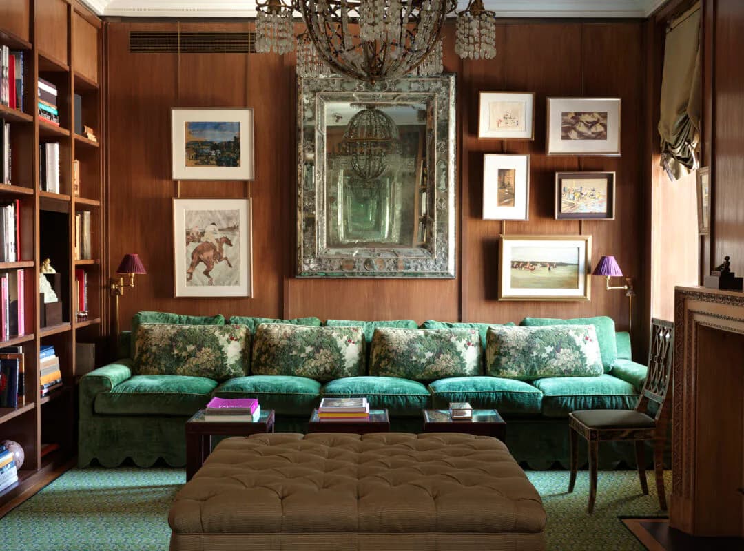 Rich library interior with cognac-brown walls, emerald velvet sectional sofa, brown ottoman, ornate mirror, and framed artwork collection by Charlotte Worthy Architects.