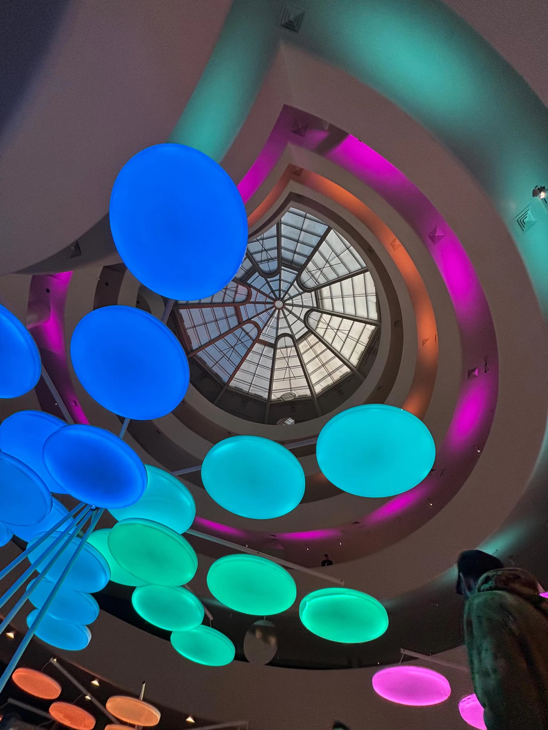 Spiral stairwell interior featuring geometric skylight, neon-lit spherical installations in blue, cyan, and magenta, and dramatic architectural curves by Chateau Brooklyn.