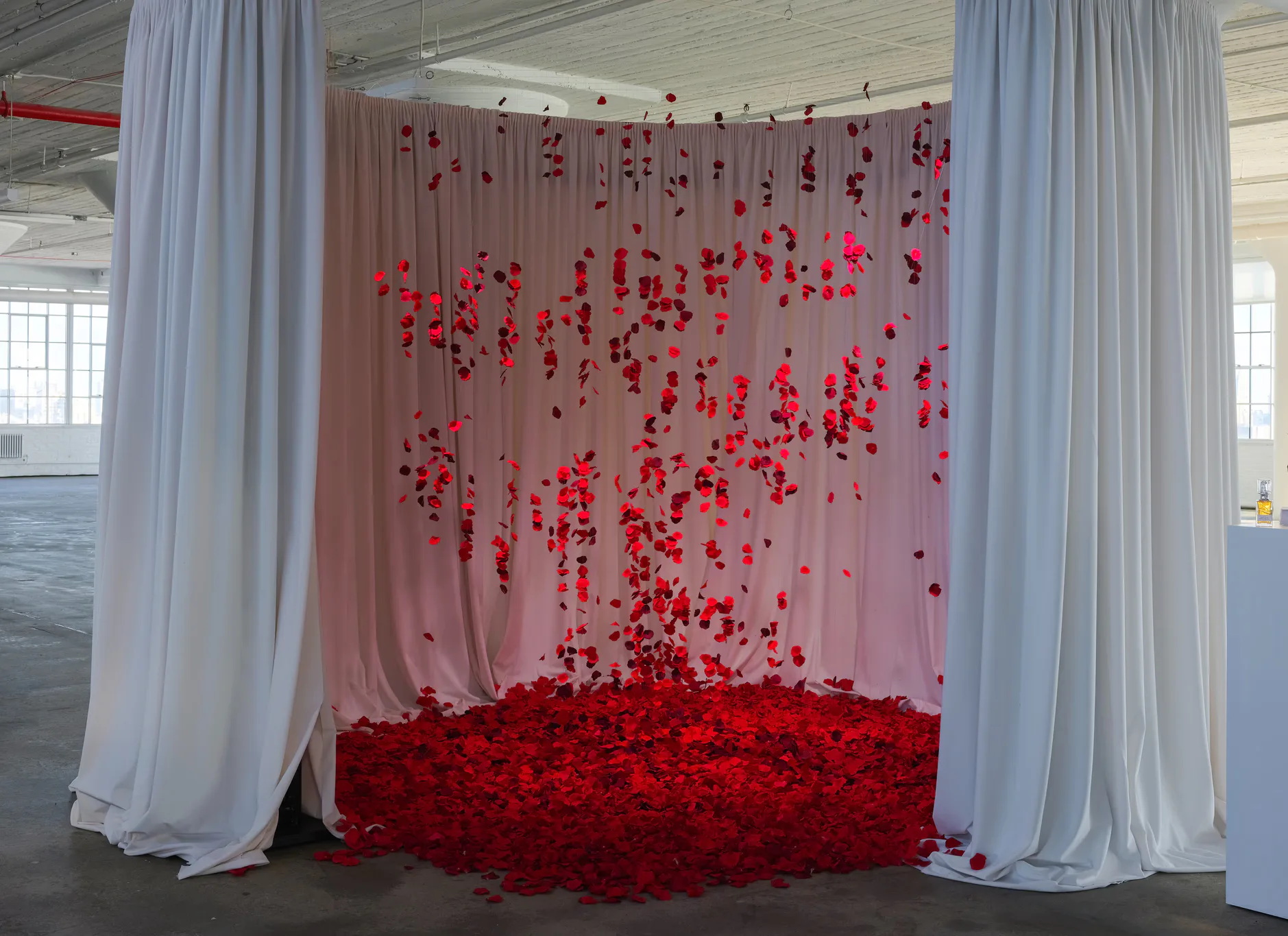 Red floral installation with suspended blooms and petals cascading onto ground, framed by white curtains in industrial space by Chateau Brooklyn.