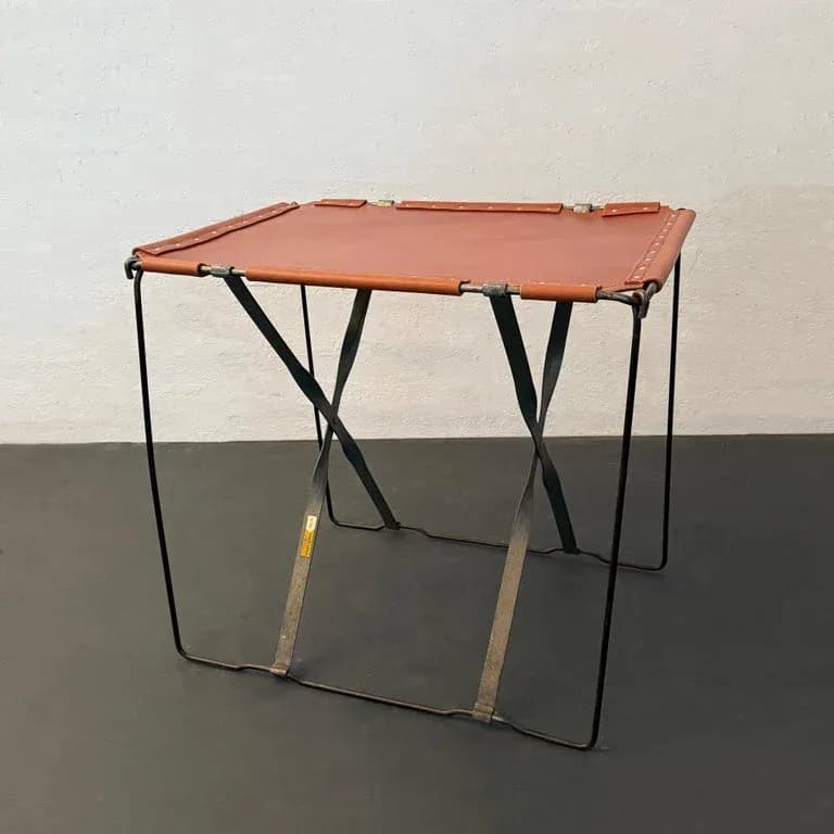 Minimalist camping table with rust-orange fabric top and black metal X-frame legs, photographed against white wall by cityFoundry.