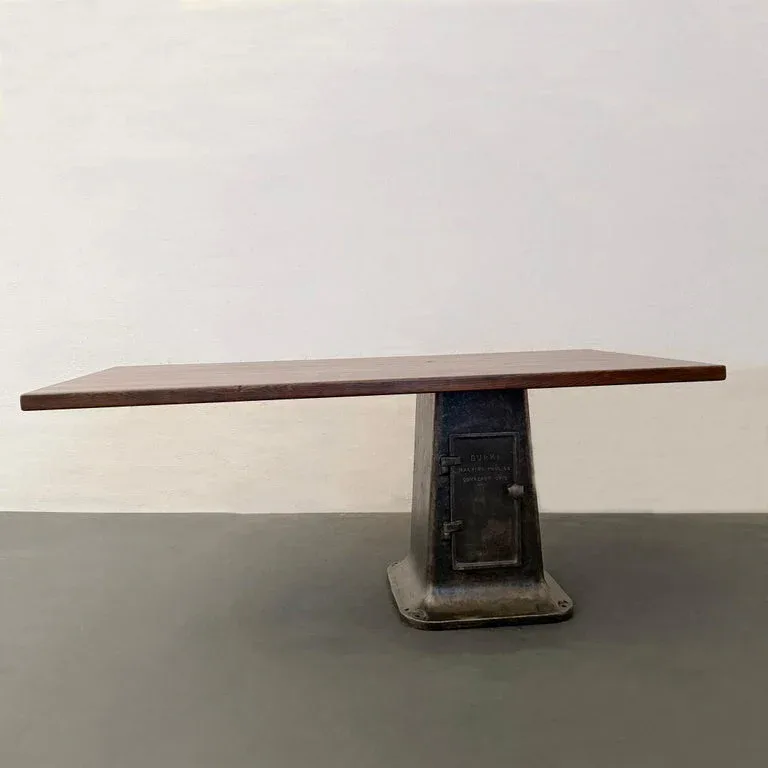 Minimalist wooden table with thin rectangular top balanced on a dark sculptural pedestal base, photographed against neutral backdrop by cityFoundry.