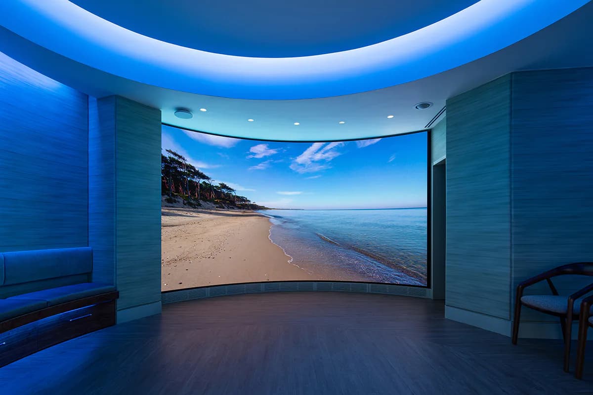 Modern curved theater space with blue ambient ceiling lighting and large curved beach landscape display screen, designed by Cline Bettridge Bernstein Lighting Design (CBB).