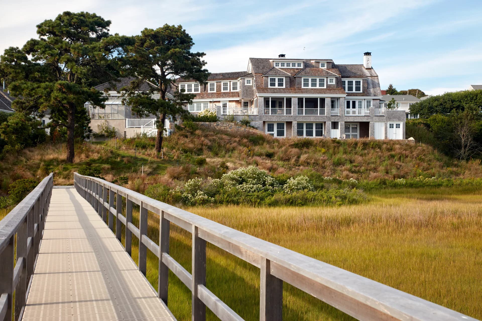 Coastal residential landscape with elevated boardwalk overlooking salt marsh, leading to multi-story shingled mansion on hillside by Clodagh Design.