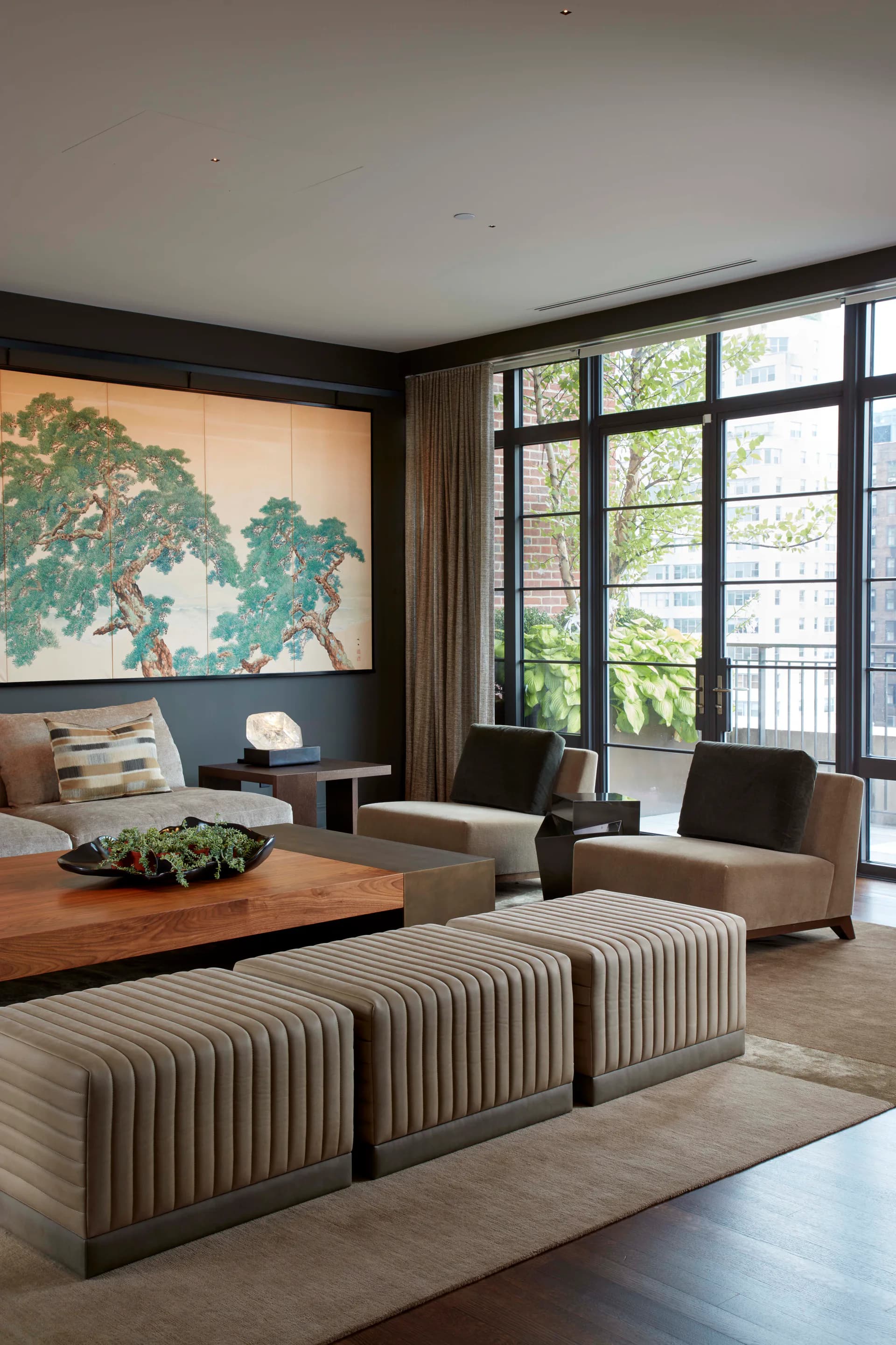 Contemporary living room with blue-gray accent wall, tree artwork, wooden coffee table, ribbed taupe ottomans, and floor-to-ceiling windows overlooking cityscape by Clodagh Design.