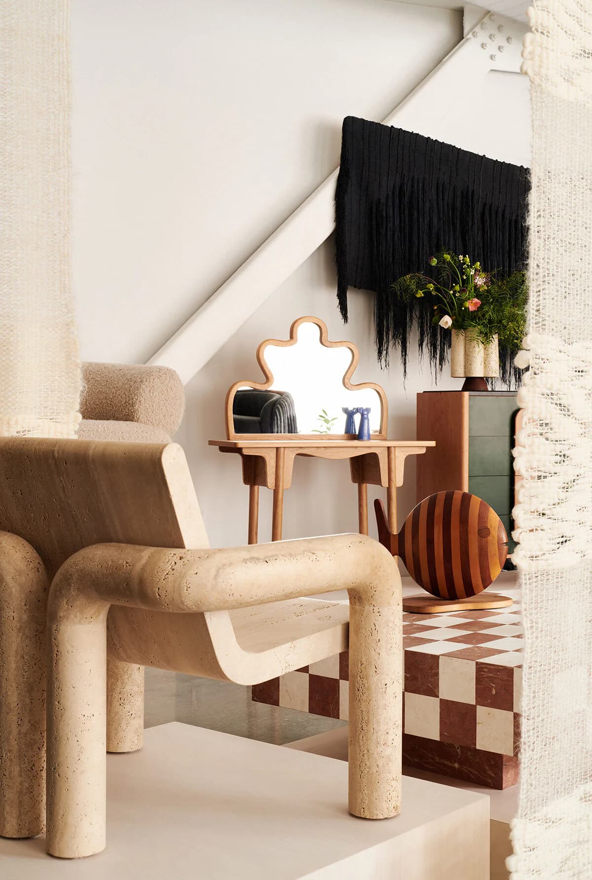 Minimalist interior with curved beige furniture, wooden puzzle-mirror, black fringed wall hanging, and geometric floor pattern by Colony.