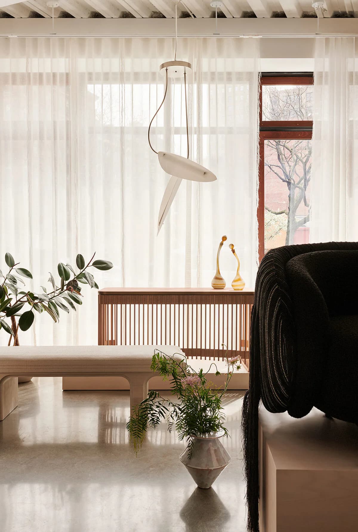 Minimalist living room with sheer curtains, wooden slat credenza, black sofa, potted plants, and sculptural pendant light by Colony.