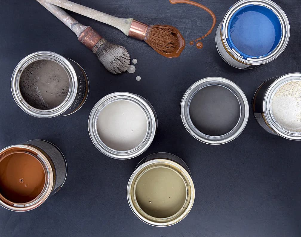 Paint color palette displaying nine open containers in shades of blue, gray, white, black, and brown, arranged with brushes on a dark surface by Color Atelier.