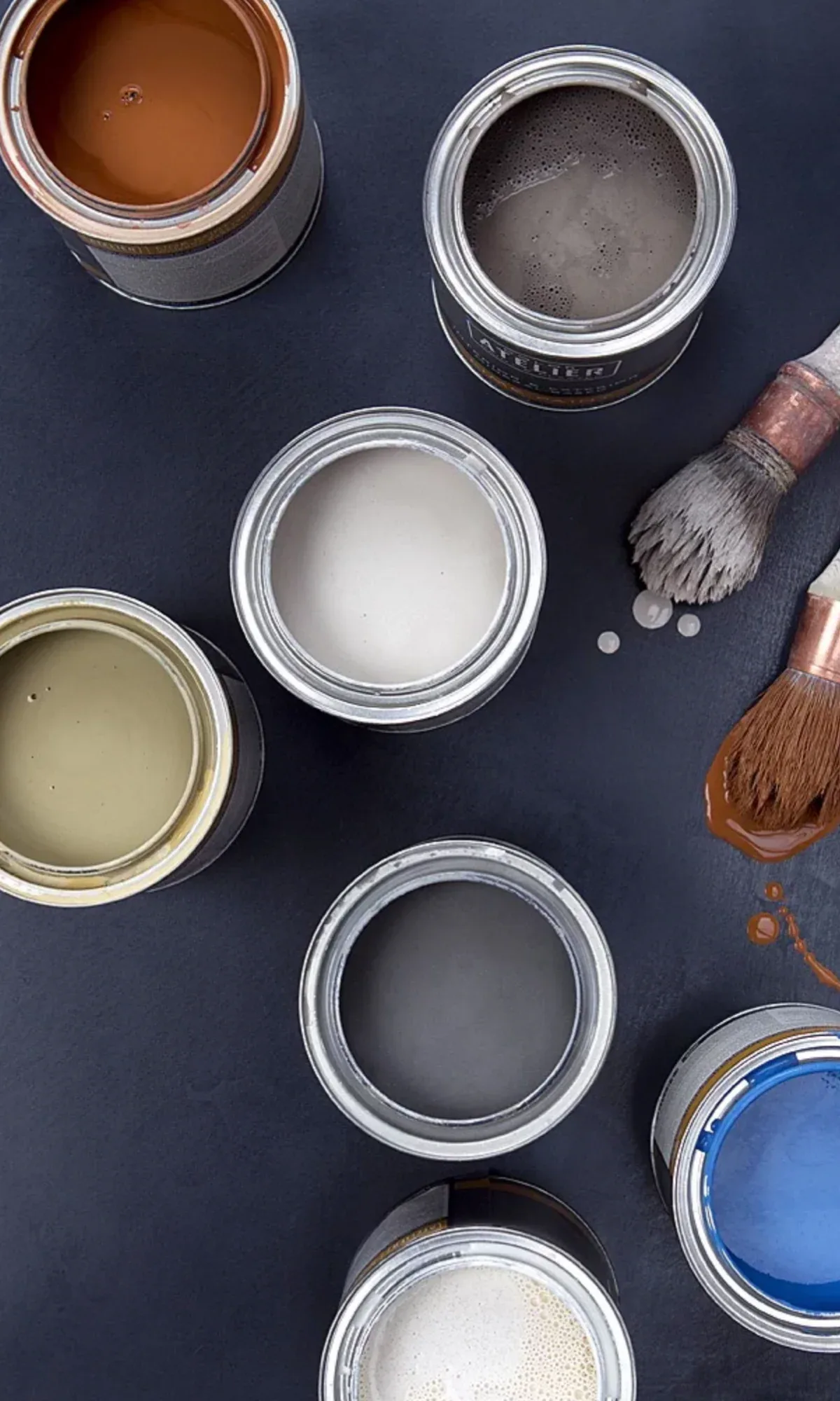 Paint color palette with open jars in neutral and warm tones arranged on dark surface with brushes by Color Atelier.