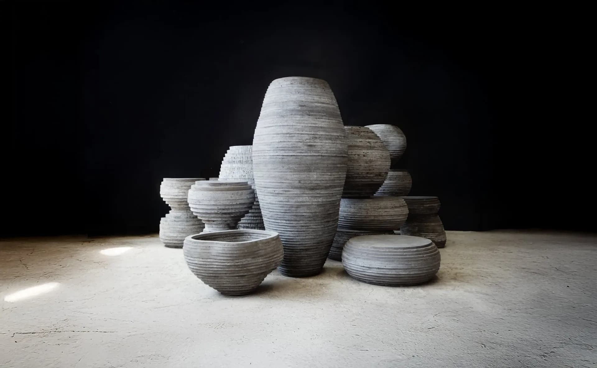 Cluster of textured ceramic vessels in varying heights with horizontal striations, arranged on a light concrete floor against a dark background by Concrete Poetics.