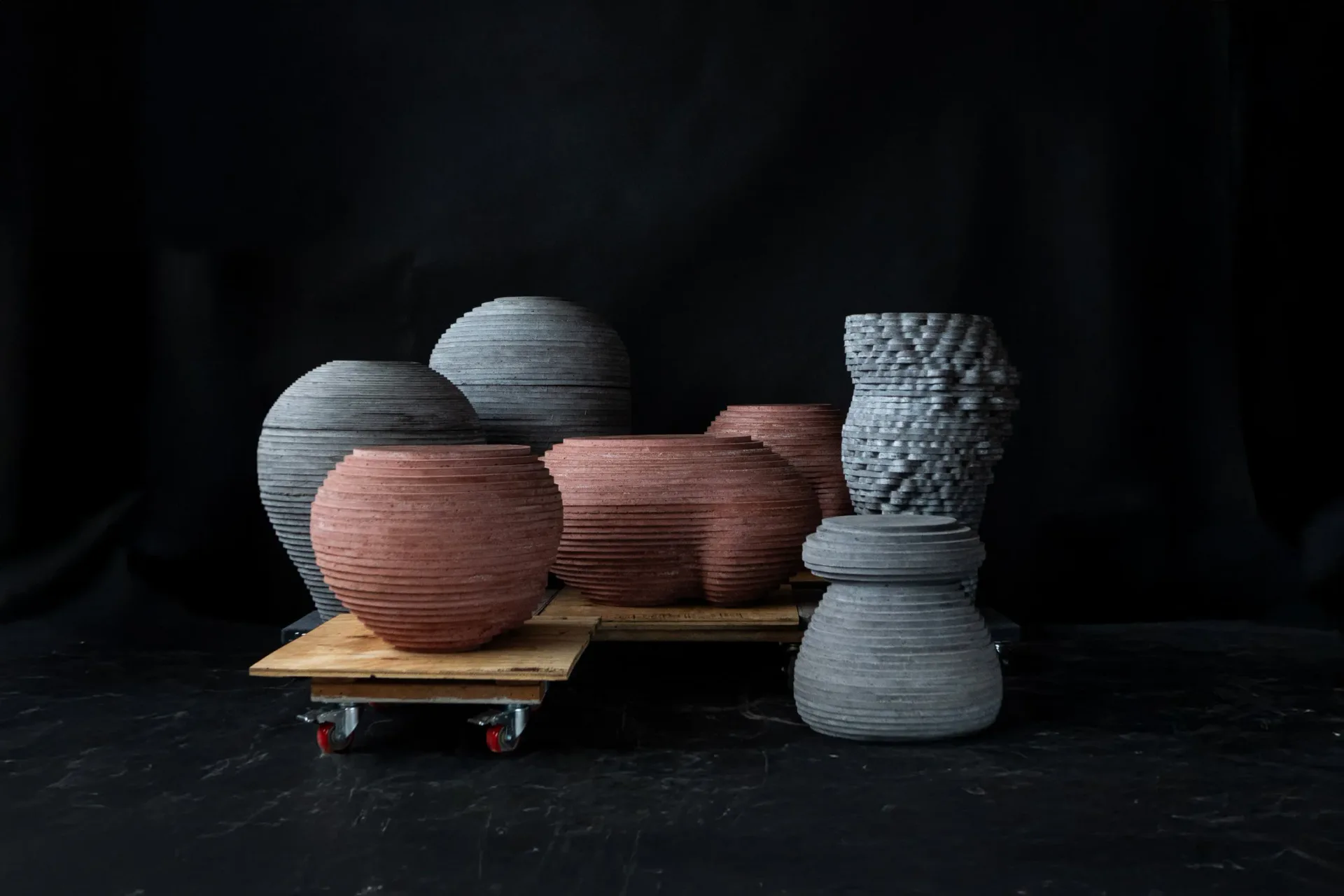 Abstract composition of textured spherical and cylindrical forms in muted earth tones and grays arranged on a wooden platform with skateboard wheels, set against a dark background by Concrete Poetics.