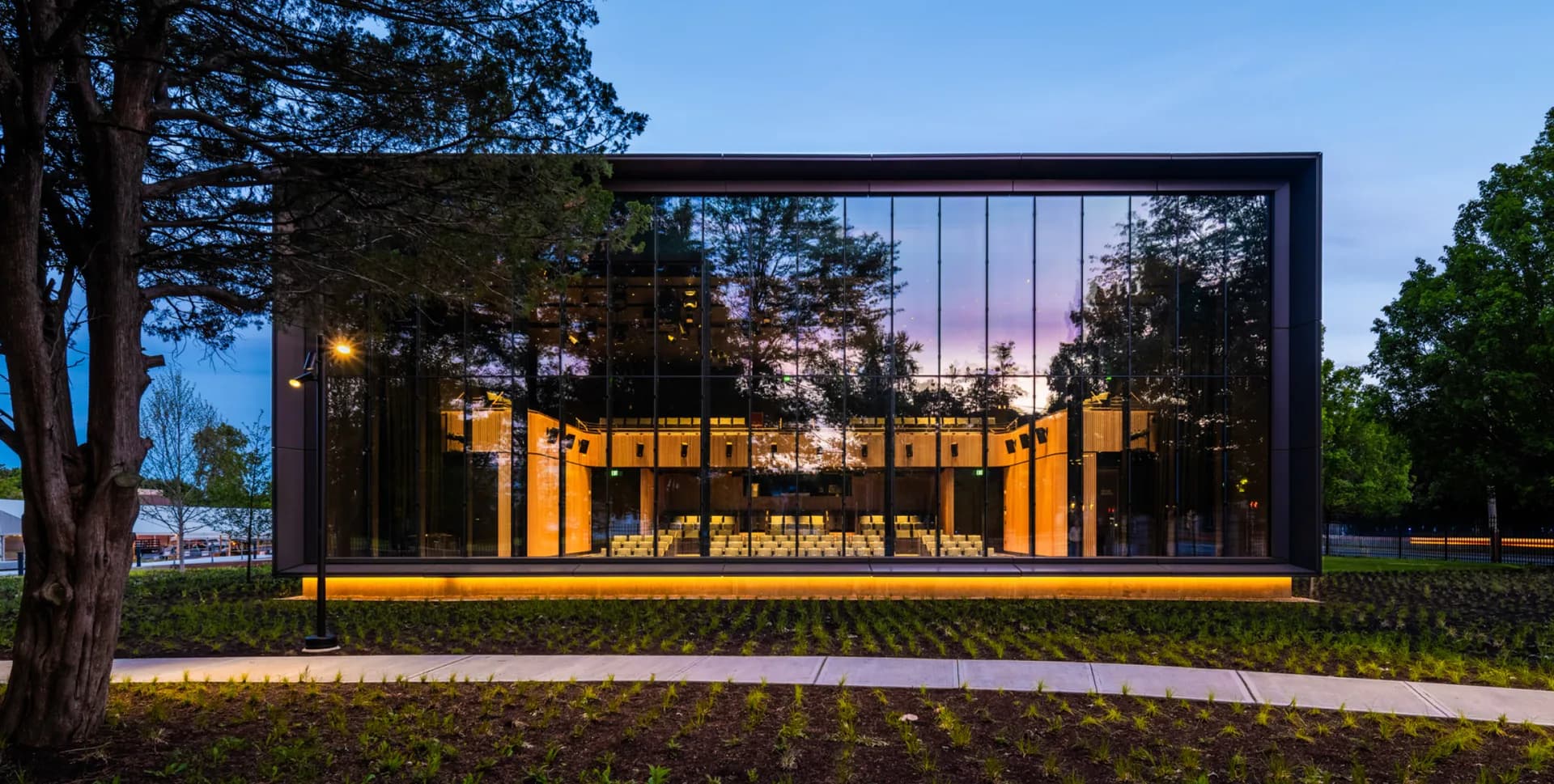Modern pavilion with black steel frame and golden-yellow interior illuminated at dusk, set among mature trees, by COOKFOX Architects.