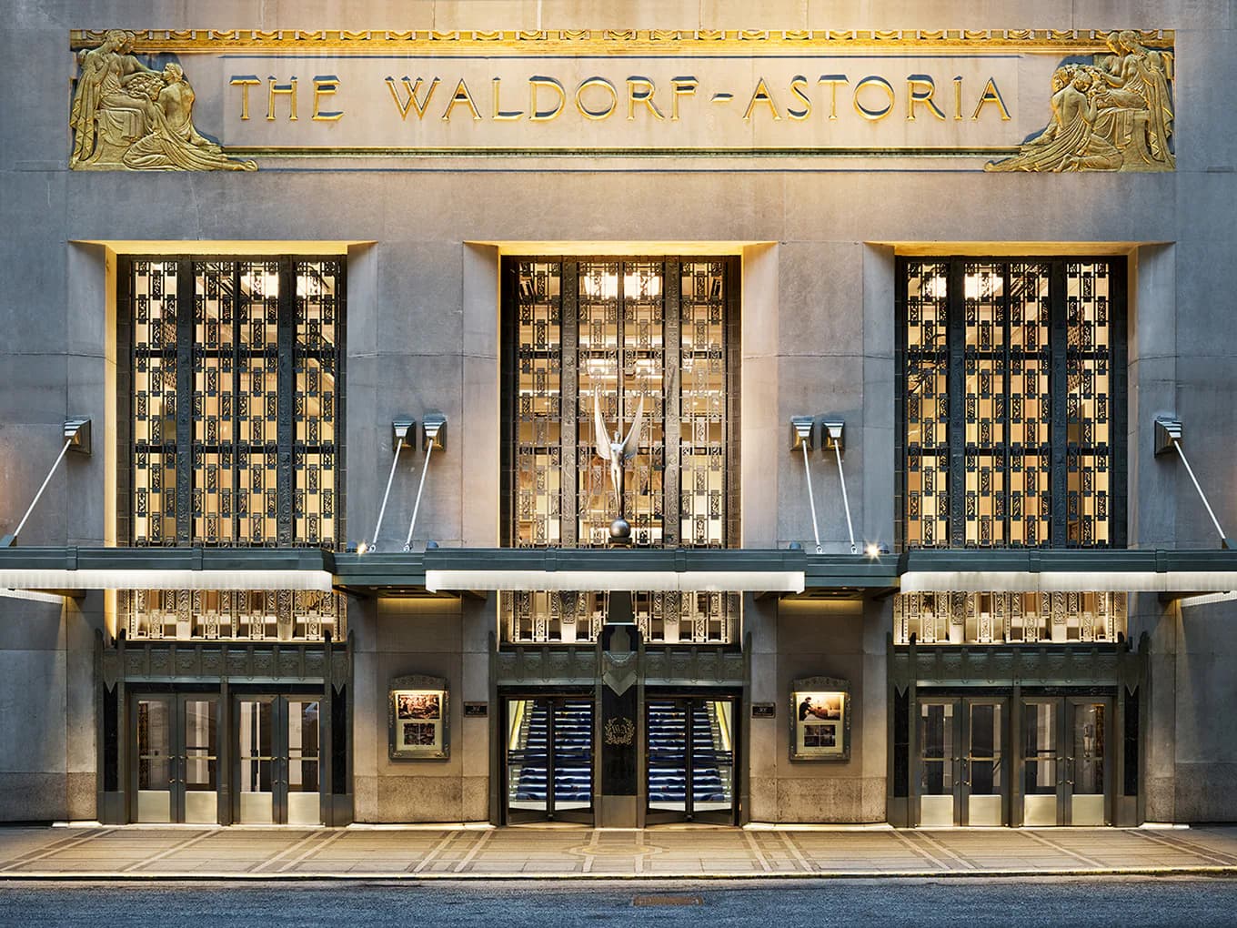 The Waldorf Astoria's Art Deco facade features three golden geometric patterned doors flanked by illuminated display windows, with ornamental metalwork and sconces by Cooley Monato Studio.