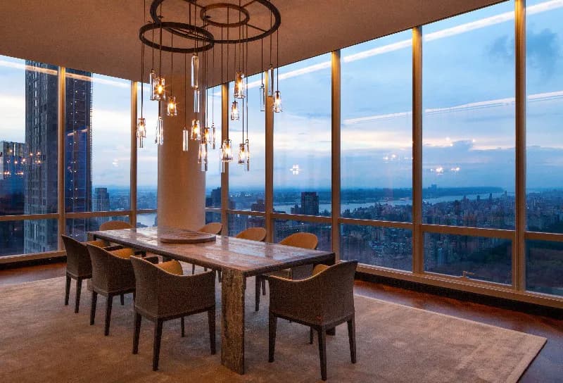 High-rise dining space with floor-to-ceiling windows overlooking a cityscape at dusk, featuring a wooden table, modern pendant lighting, and warm wood framing by Cooley Monato Studio.